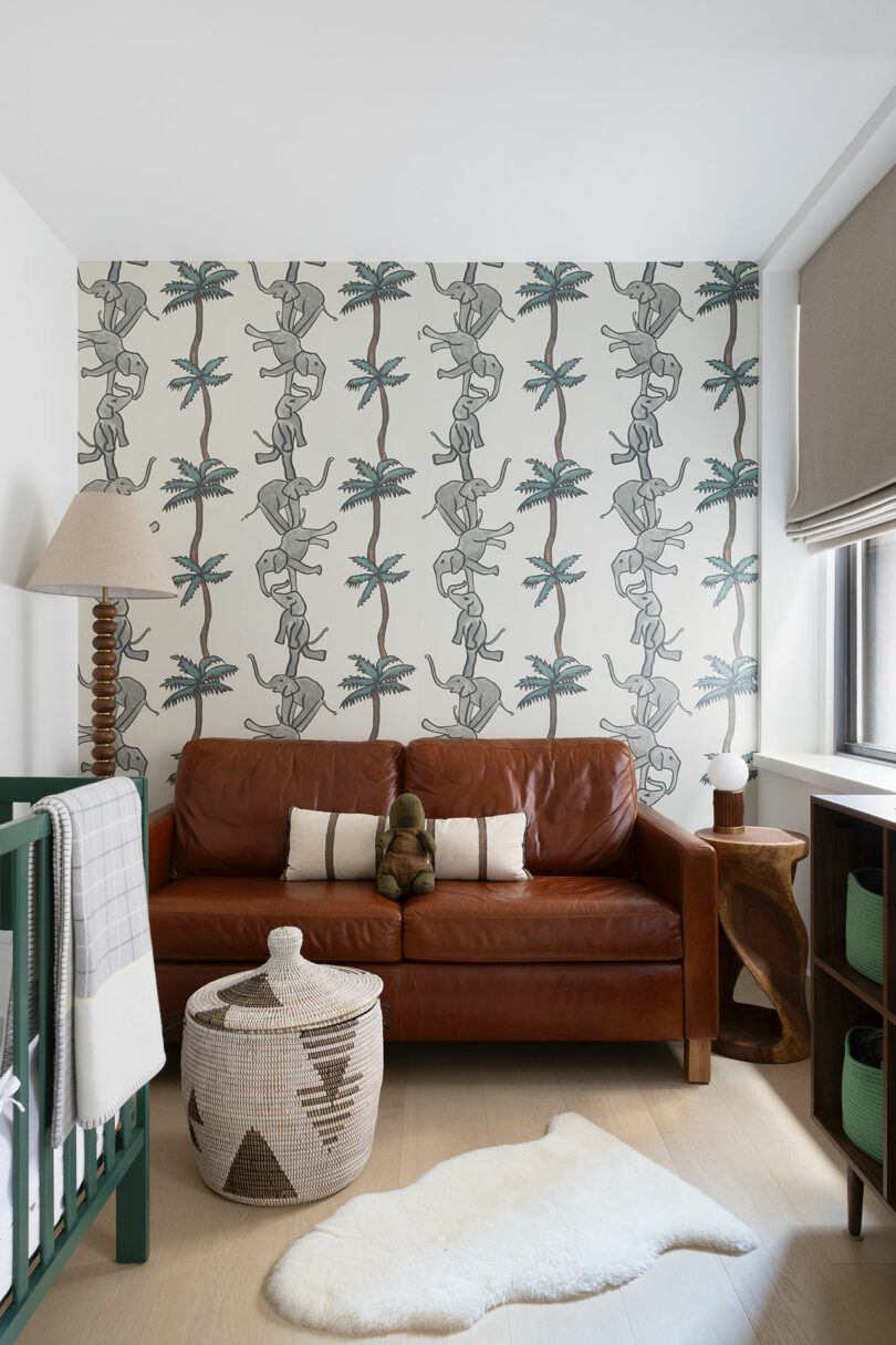 A cozy den features a brown leather sofa, a crib, a woven basket and palm-themed wallpaper.  A plush toy sits on the sofa and a sheepskin rug lies on the wooden floor.